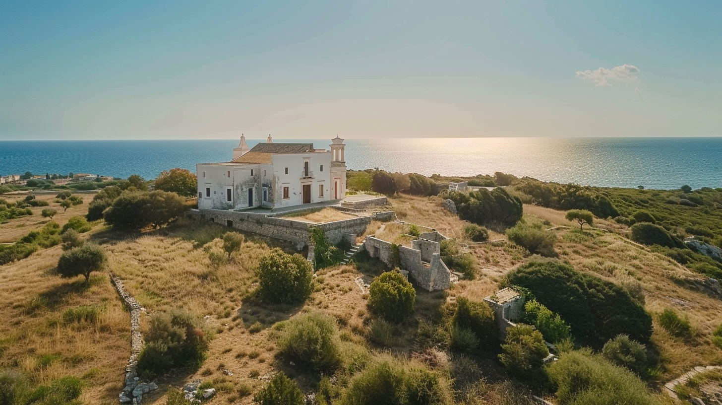 Una grande villa storica bianca con dettagli architettonici eleganti, situata su un terreno elevato, circondata da vegetazione secca e con vista sul mare azzurro.